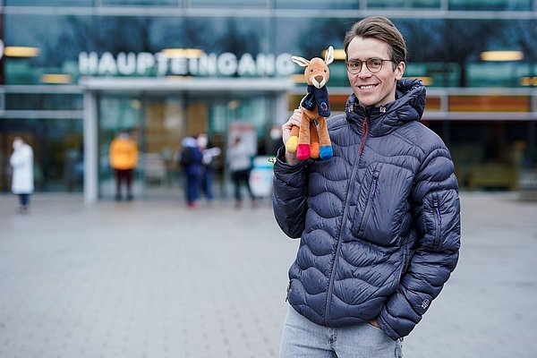 ohannes Wimmer vor Eingang des Zentrums für Kinder- und Jugendmedizin des UKHD mit KiTZ-Maskottchen „Hopps“